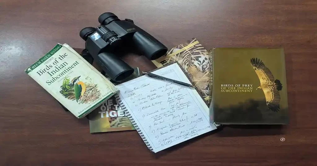 The image represents few things we require for birding, namely a pair of good bino, a birding guide book, a diary and a pen . 